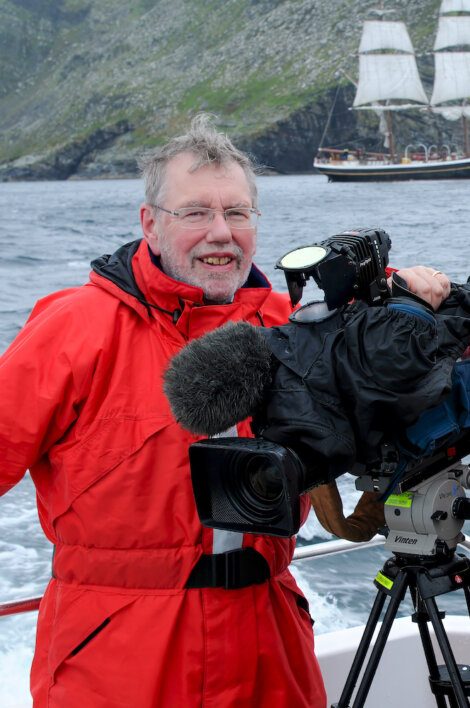 Man in red jacket operating a video camera on a boat, with a tall ship in the background on a cloudy day.