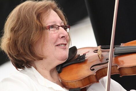 A smiling woman playing a violin, wearing glasses and a white shirt, focused intently on her performance.