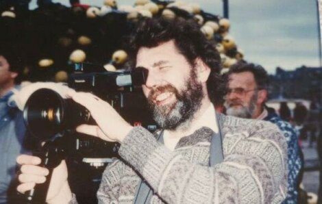 A man with a beard smiling while taking a photo with a professional camera, outdoors during the day with people in the background.