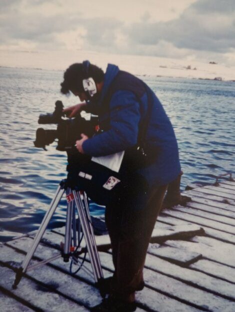 A person in a clown mask operates a camera on a tripod on a wooden pier with a snowy landscape and water in the background.