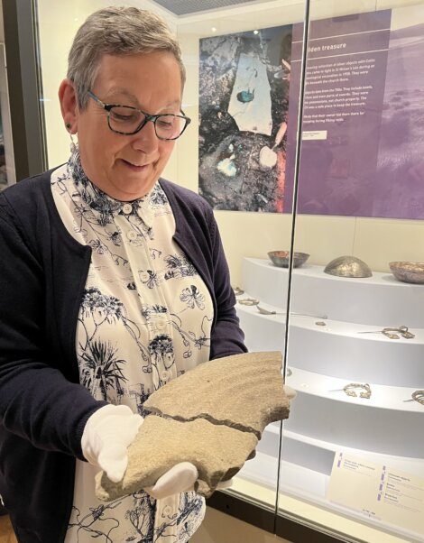 A woman examining an ancient pottery shard in a museum exhibit, wearing gloves and smiling, with informational displays in the background.