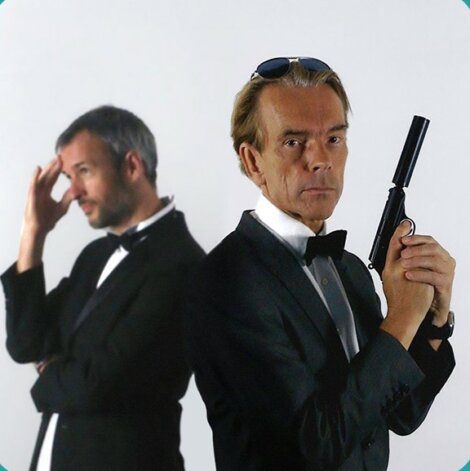 Two men in tuxedos, one holding a gun and wearing sunglasses, the other looking pensive with his hand on his forehead.