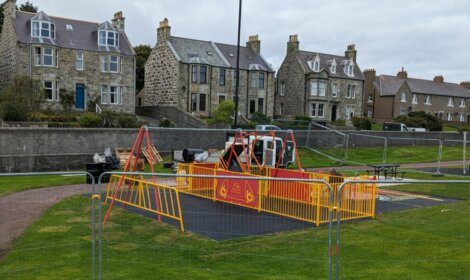 A playground with equipment cordoned off by yellow safety barriers, with traditional stone houses in the background.