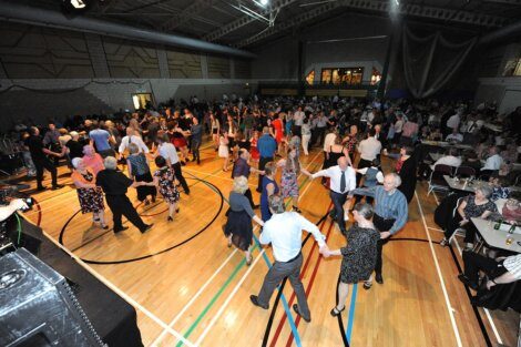 People dancing and socializing at an indoor event or gathering with tables set around the perimeter of the dance floor.