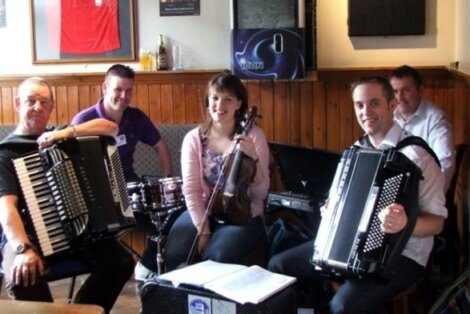 Group of musicians with accordions and a violin smiling for the camera in a pub setting.