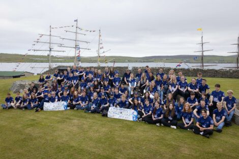 A large group of individuals wearing matching blue shirts gathers for a group photo in front of a tall ship docked at a grassy waterfront.