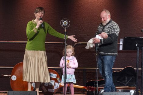 A woman speaking into a microphone on stage with a man holding a baby next to a young child amidst musical instruments.
