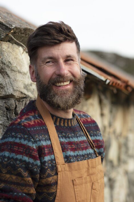 Man with a beard smiling outdoors while wearing a patterned sweater and overalls.
