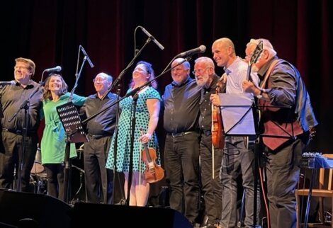 A group of musicians with various instruments standing on stage, smiling and taking a bow after a performance.