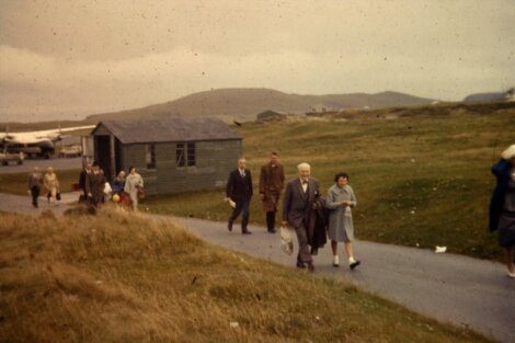 Elderly couple walking on a path with others in a grassy area, with a wooden hut and hills in the background.