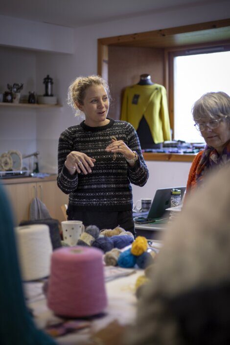 Woman explaining or demonstrating something during a workshop with yarn and knitting materials on the table.