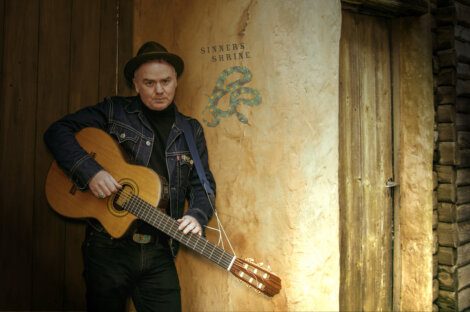 A man leaning against a wall with an acoustic guitar.