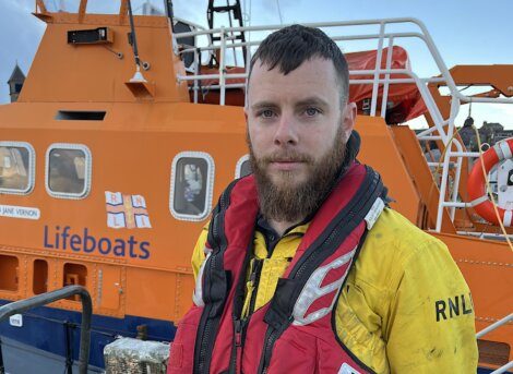 A man in a lifeboat stands in front of an orange boat.