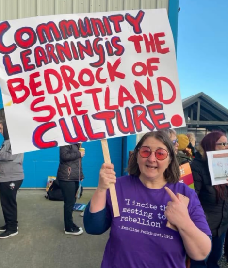 A woman holding a sign that says community learning the bedrock of shetland culture.