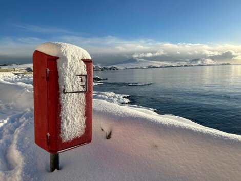 A red post box covered in snow near the ocean.