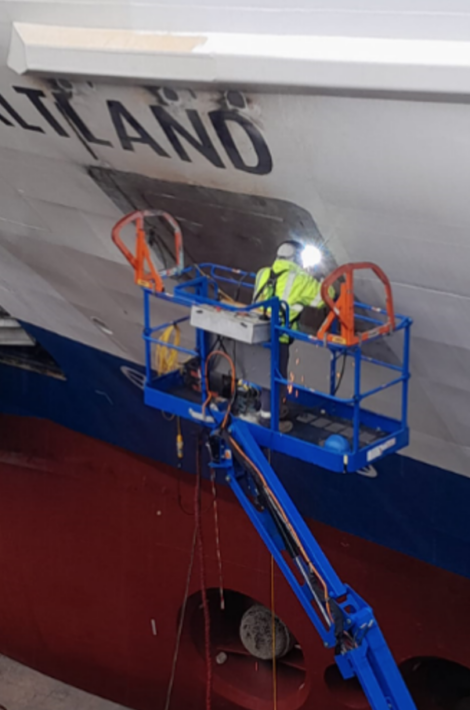 A worker in high-visibility clothing is operating a crane-mounted elevated work platform to perform maintenance or repairs on the hull of a ship named “atland.”.