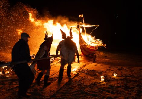 Individuals dressed in viking attire pushing a burning boat during a nighttime ceremony.