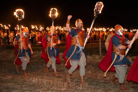 Participants in historical reenactment dressed as viking warriors march with torches at night.