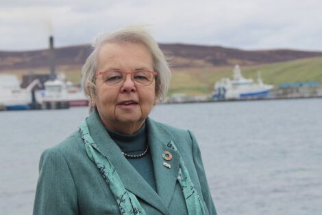 An elderly woman with glasses, wearing a green scarf and blazer, standing in front of a harbor.