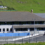 Modern school building with a blue fence in a green hilly landscape.
