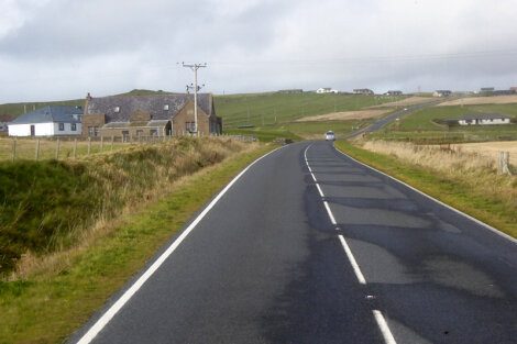 An empty country road leading towards a small collection of buildings with hills in the distance under a cloudy sky.