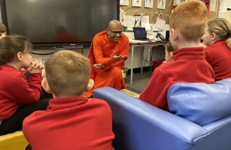 A monk in orange robes interacting with a group of schoolchildren sitting around him.
