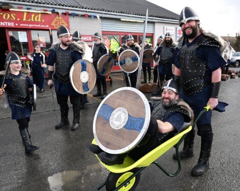Adults and children dressed as vikings, one of whom is pushing another in a wheelbarrow, enjoying a festive event.