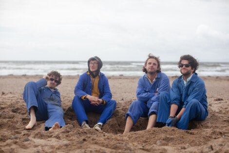 Four men in coveralls sitting on a sandy beach.