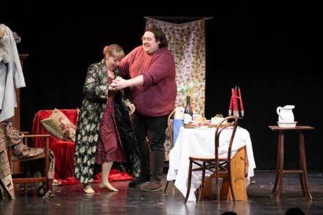 Two actors on stage performing a scene with one helping the other with their coat.