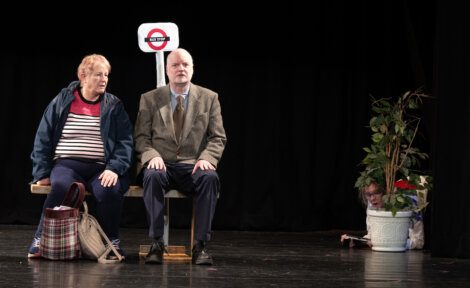 Two actors seated on stage with a third actor peeking from behind a potted plant, in a scene resembling a public transportation setting.