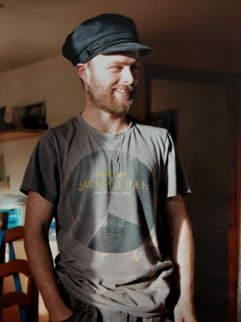 A smiling man in a flat cap and graphic t-shirt stands indoors, bathed in sunlight, looking off to the side.