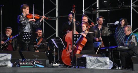 A diverse group of musicians performing on a stage, including violinists, a cellist, and guitarists, with music stands and microphones.
