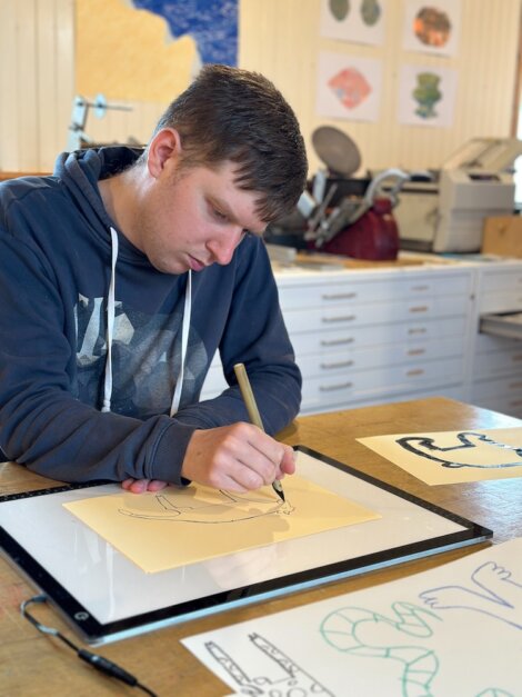 Young man concentrating on drawing with a pen on yellow paper in an art studio with sketches and supplies around.