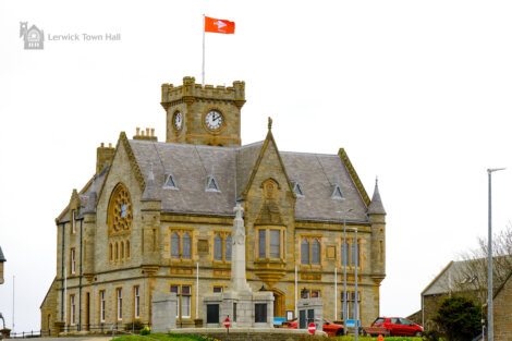 Lerwick town hall in shetland with a red flag fluttering atop, featuring a stone facade and clock tower against a cloudy sky.