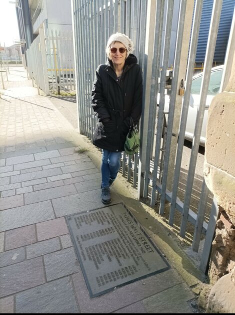 Woman in sunglasses and a black coat standing on a sidewalk by a metal fence, near a historical marker plaque embedded in the ground.