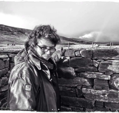 A person wearing glasses and a jacket stands beside a stone wall in a windswept landscape.