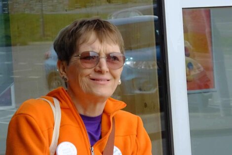 A smiling elderly woman wearing sunglasses and an orange jacket, sitting outdoors with a reflective window in the background.