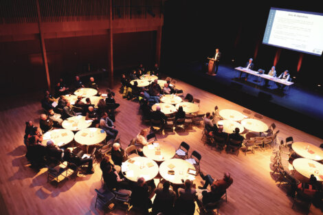 A conference hall with attendees seated at round tables, listening to a speaker at a podium with a panel seated beside a large presentation screen.
