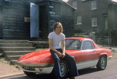 Man sitting on the hood of a red lotus elan sprint sports car, parked in front of a rustic building and garage labeled 
