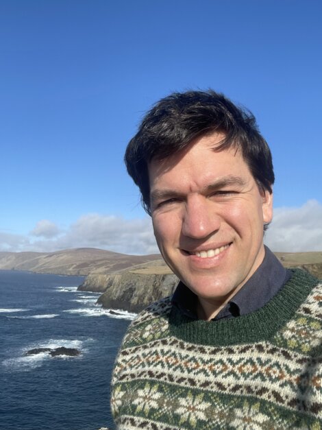 A person smiling for a selfie with a coastal landscape in the background.
