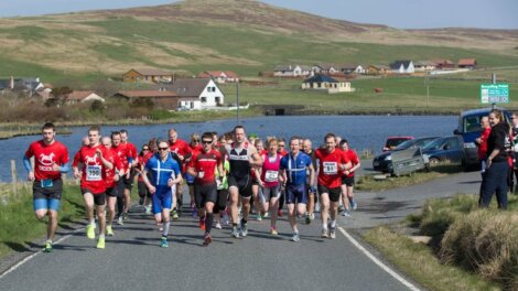 Group of runners competing in a marathon on a rural road, with houses and hills in the background.